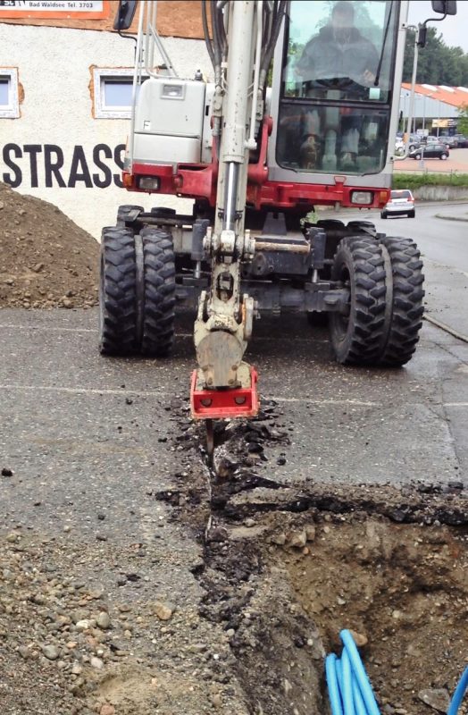 Mit der Kante der Wurzelratte schneidet der Baggerführer zunächst V-förmig,etwa 12 bis 15 Zentimeter tief und mehrere Meter lang, in den Asphalt.